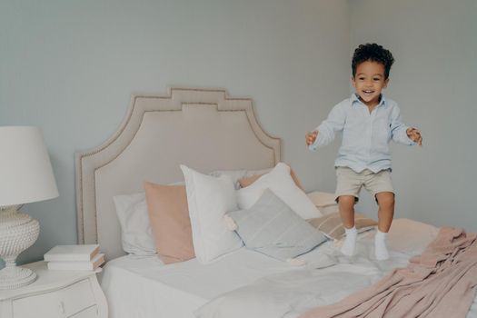 Ecxited mixed race ethnicity kid boy in casual clothes having fun and jumping on big bed, with white and pink beddings and pillows. Photo taken in air movition with child playing indoors