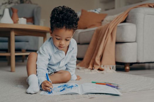 Small focused african baby boy sitting on comfortable carpeted floor in living room, drawing with blue pencil in coloring book. Children leisure time at home and creative hobbies for kids concept