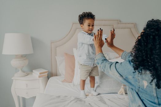 Cute small afro american kid playing engaging games on big bed in light themed bedroom with his caring curly mother, holding their hands together. Loving motherhood and child care concept