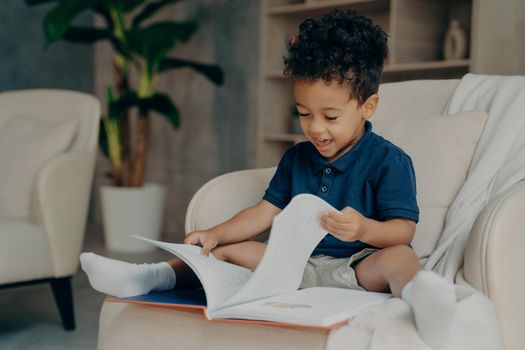 Happy mixed race little kid boy with cute curly hair reading story book while sitting in cozy armchair in light colored living room at home, child spending free time indoors. Children development