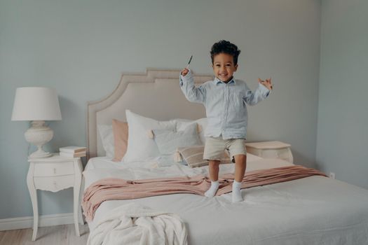Joyful small mixed race boy happy kid dressed in blue shirt and beige shorts jumping on big comfy bed with candy, expressing his happiness with shining smile, enjoying playing alone at home