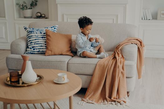 Photo of living room with small lovely mulatto child peacefully sitting on sofa among pillows and coverlid in foreground of coffee table with teacup and vases decorations, kid playing at home