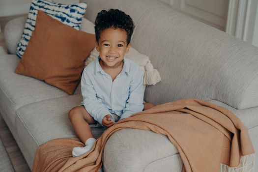Lovely afro american child with curly hair and cute smile sitting on sofa with beige color elements pillows and coverlid, looking up on camera and smiling while playing in living room at home