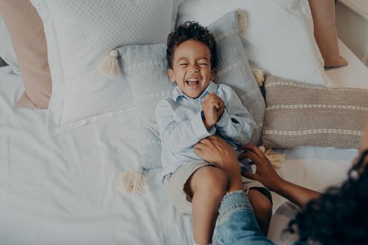 Woman mom tickles adorable mixed race little son with curly hair lying on pillows in bed. Cute baby boy in light colored casual clothes having fun and sincerely laughing. Family weekend leisure time