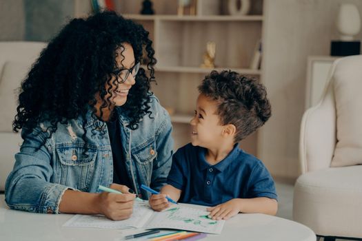 Beautiful afro american family of two, mother and son looking at each other while spending time at home by painting book with different colors in living room. Motherhood and parenting concept