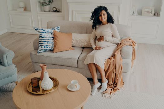 Young african pregnant woman in casual dress caressing her belly while sitting on cozy couch in stylish living room ambiance. Happy future mom expecting baby and maternity leisure time at home concept
