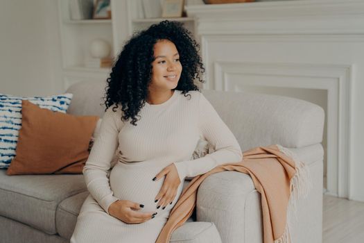 Beautiful young pregnant woman hugging her tummy while relaxing on couch at home, millennial expectant mom resting on sofa in living room and smiling, enjoying pregnancy and future maternity concept