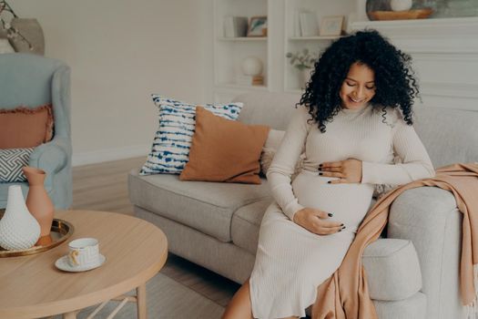 Smiling lovely pregnant mixed race woman in white dress with curly hair sitting on couch in front of round table and thinking about motherhood, happy to become mother. Pregnancy concept