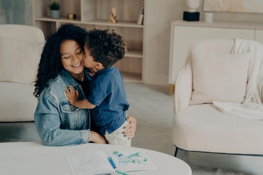 Cute mixed race boy little kid gently hugs his happy mother sitting on floor by table with colored pencils on it, surrounded by light colored furniture in living room. Leisure time with family concept