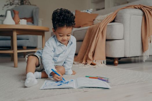 Lovely african child sitting on floor at home in stylish living room with colored felt tip pens and drawing in album with focused expression on his face while spending leisure time at home
