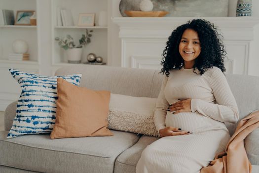 Smiling pretty expectant young mom with curly hair elegantly hugging belly and looking gladfully at camera, sitting on sofa full of pillows against white wall in modern living room, enjoying pregnancy