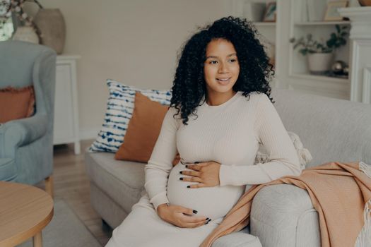 Smiling lovely african expectant young mom sitting on sofa with colorful pillows tenderly holding belly, against white wall background in modern living room in apartment interior. Pregnancy concept