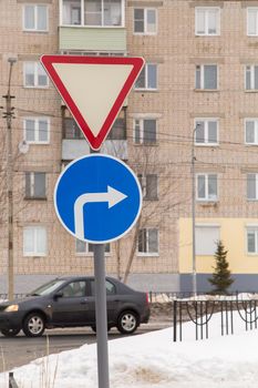 Traffic signs mean give way and turn right only. Against the background of a road and a residential building. It's winter in the city, white snow lies on the street.