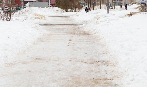 Winter sidewalk in the city, soft snow covers the path going into the distance. On the road you can see the thawing dirty snow. In the background, in the distance, houses, people, trees, a car.