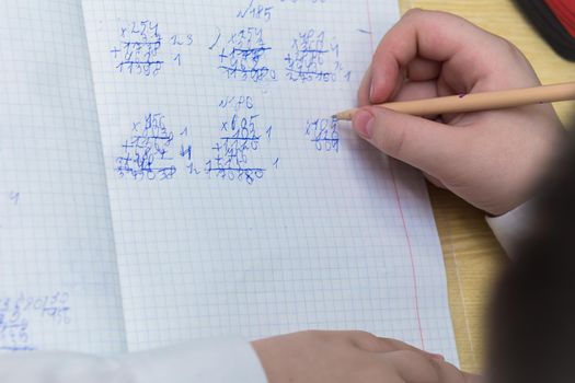 A girl's hand with a ballpoint pen close-up writes in bad handwriting. The student completes the task. A student solves examples in mathematics at school at a desk. Warm soft daylight.
