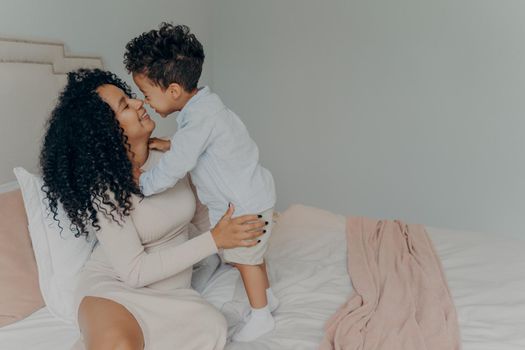 Mixed race happy pregnant woman mother embracing adorable baby boy while sitting relaxing on bed in bedroom at home. Sweet motherhood moments of maternity and joy of being mom concept