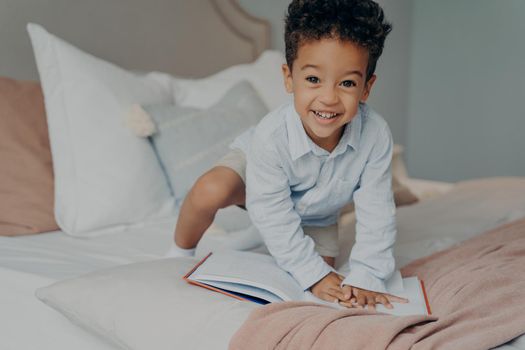 Cute photo of small afro american smiling child in light blue shirt sitting on parents bed with story book early in morning on weekend. Happy boy kid spending leisure activity time at home concept