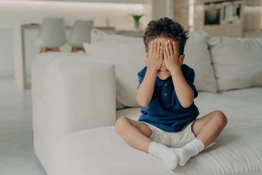 Small excited boy in tshirts and shorts playing game of hide and seek, covering face with his hands, eager to start searching, waiting and counting while sitting on white couch in light modern room
