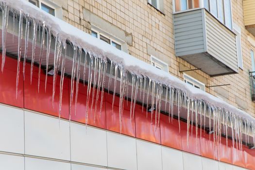 Long, even icicles hang from the roof against the backdrop of the house. The wall of the building is finished with red and white plastic tiles. Abnormal weather concept. New Year and Christmas is over