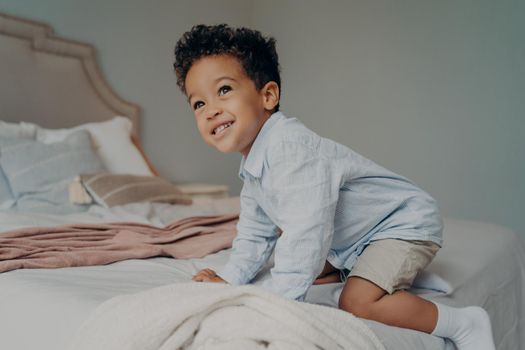 Cute smiling mixed race child toddler with black curly hair tries to climb on big bed of his parents in light shirt and shorts on weekend morning. Small kid having fun, enjoying and playing indoors