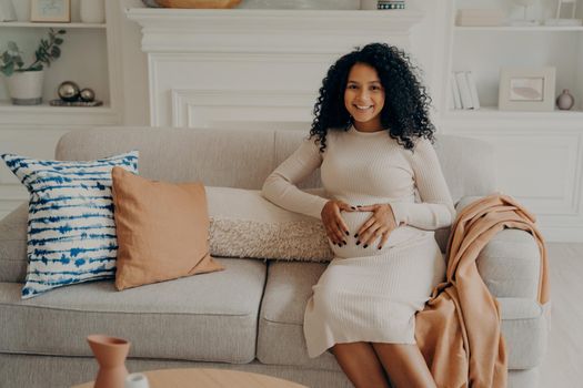 Women and maternity concept. Portrait of lovely sweet expectant mixed race lady sitting on soft couch decorated with cushions with her hands folded on belly in form of heart. Happiness of pregnancy