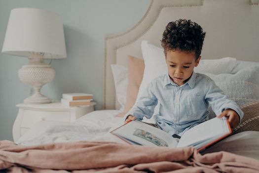 Cute focused small boy in casual wear holds and tries to read childrens book, sitting on bed with nightstand next to with white elegant lamp on it. Kid spending weekend morning leisure at home