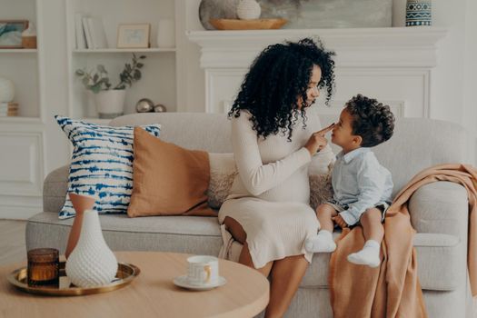 Pregnant mother and son afro ethnic race sitting on sofa in modern living room interior, mom talking to boy touching with finger tip of his nose, child listens very attentively. Motherhood concept