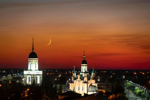 A church,temple or cathedral against the background of an evening sunset with a maroon sky and a big month.The horizon line at dawn with the moon and the red sky.City panorama with towers and domes