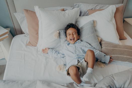 Top view of mixed race laughing and having fun little boy lying on pillows on bed in parents bedroom in weekend morning wearing casual clothes, happy kid spending leisure time indoor