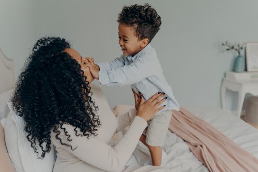 Happy afro american family, pregnant woman loving mom in white dress with curly hair sitting on white bed and joyfully spending time with child, giving attention to her son and playing with him
