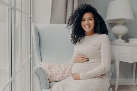 Afro American adorable smiling pregnant lady in knitted beige dress peacefuly sits in white armchair next to large light window placed hands on her belly at home. Enjoying maternity time concept