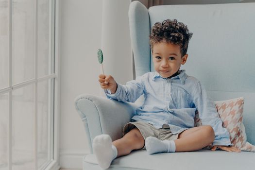Portrait of cute adorable little mulatto boy with big eyes holding round blue sweet and tasty lollipop and looking at camera, casually dressed sitting alone on comfy armchair in minimalist room