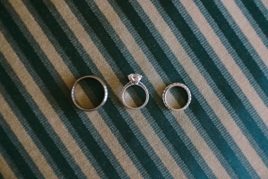 Close-up of white gold wedding rings and a gemstone engagement ring for a bride on blue fabric with white stripes. High quality photo