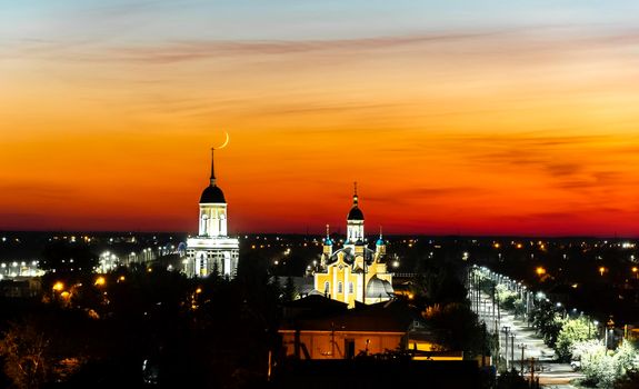 A church,temple or cathedral against the background of an evening sunset with a maroon sky and a big month.The horizon line at dawn with the moon and the red sky.City panorama with towers and domes