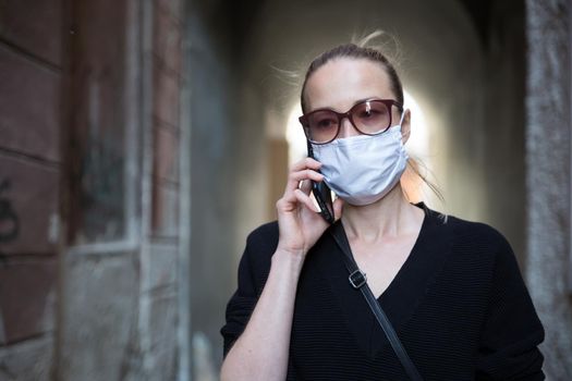 COVID-19 pandemic coronavirus. Casual caucasian woman at medieval city street using mobile phone, wearing protective face mask against spreading of coronavirus and disease transmission.