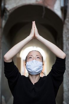 Coronavirus outbreak. Young caucasian woman wearing medical protection face mask praying over coronavirus global pandemic, for salvation of humanity, health, anxiety and depression reduction...