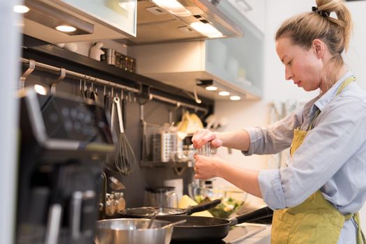 Stay at home housewife woman cooking in kitchen, salting dish in a saucepan, preparing food for family dinner. Blonde caucasian model in her 30is, real cooking not looking in camera.