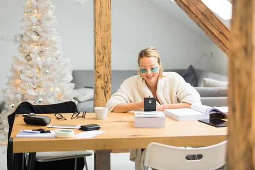 Young businesswoman wearing cosy warm bathrobe and cosmetic eye patches working remotly from home in winter Christmas time during 2020 corona virus pandemic. Work from home, selter in place, concept.