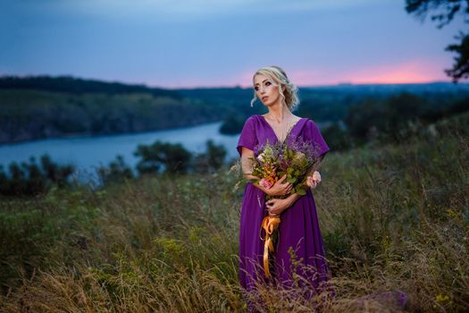 Girl model blonde in a lilac dress with a bouquet with a green forest