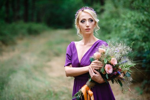 Girl model blonde in a lilac dress with a bouquet with a green forest