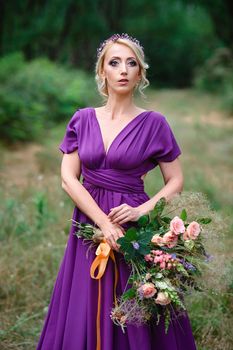 Girl model blonde in a lilac dress with a bouquet with a green forest