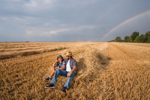 Father and son are resting after successful harvest. Rainbow in the sky behind them.