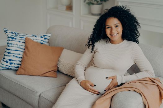Happy pregnancy time. Cheerful young expectant lovely mixed race young mother with natural looking curly hair relaxing on sofa in living room, touching belly and smiling on camera