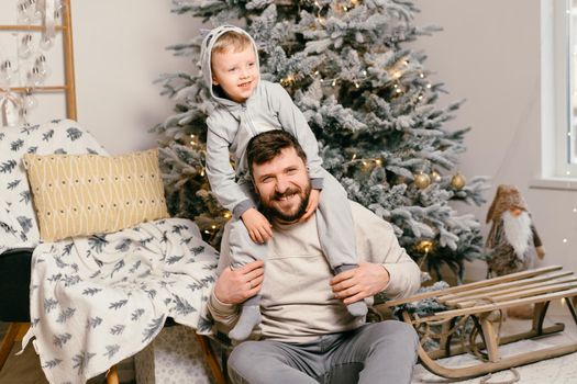 Holiday Christmas Handsome father playing with small cute little boy near decorated New year tree at home Family tradition positive son sitting on dad shoulders