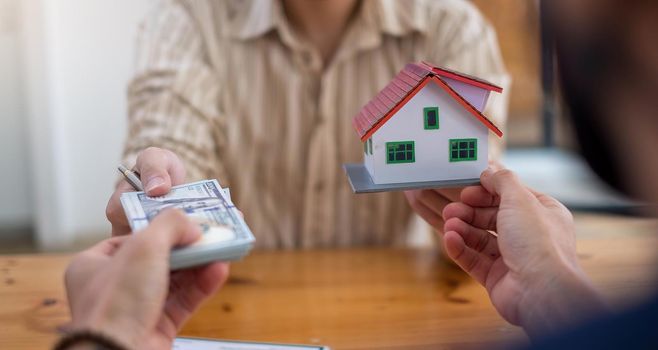 business people handing over dollars with decorative house model and contract at office.