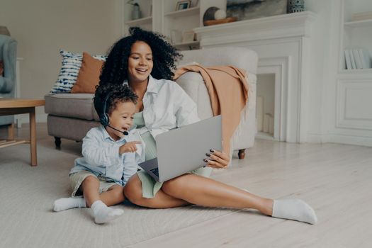 Cute mulatto little boy son wearing headset pointing at laptop while having video call with father or grandparents, enjoying online talk with family while sitting on floor with beautiful positive mom