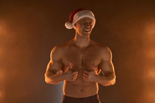 Portrait of muscular man wearing Christmas Santa hat, showing thumb up and smile on smoky background Macho Shirtless Naked torso stripper Hot sexy Santa guy Seductive male for holiday