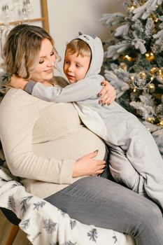 Mom plays with child. Happy family Portrait In Home pregnant mother embraces his little son. Happy new year. decorated Christmas tree Christmas morning bright living room Caucasian woman with child