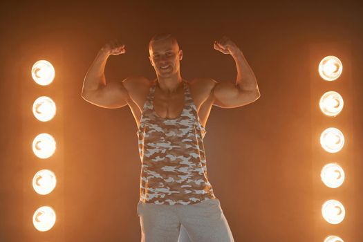 Active lifestyle concept. Professional bodybuilder showing biceps and smiling at camera, lamps illumination and smoke on background