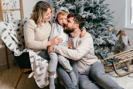 Christmas Family Happiness Portrait of dad, pregnant mom and little son sitting armchair at home near Christmas tree hug smile European young adult family holiday morning